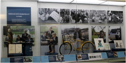 he Americans with Disabilities Act, 1990-2015 artifact wall, showing large graphics of the signing of the law by Pres. Bush, a yellow bike, and other artifacts behind glass on display in a shallow, tall case
